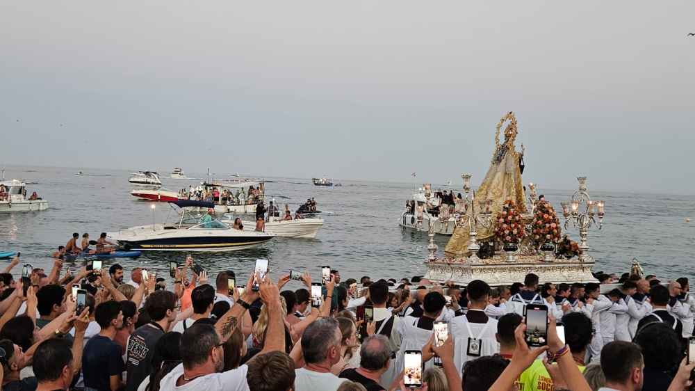 Procession with a statue by the sea, people recording with phones, and boats in the background.
