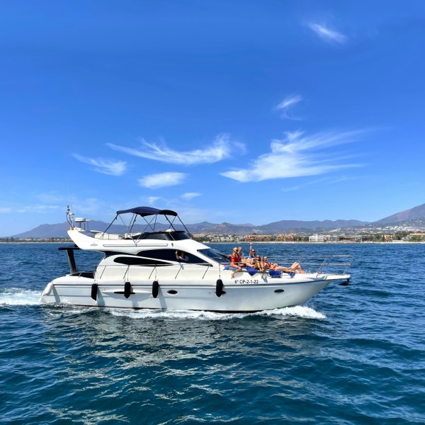 People relaxing on a yacht in the ocean with mountains in the background under a clear blue sky.