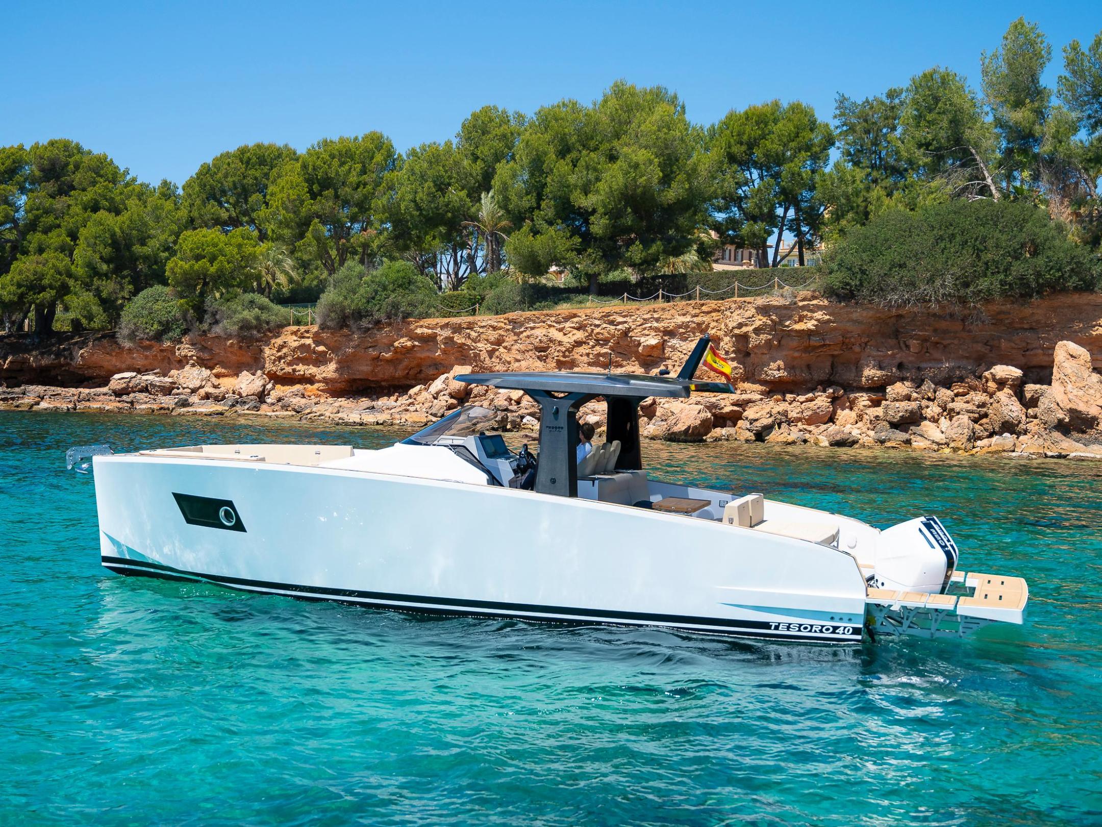 A modern white boat sails on clear turquoise water near a rocky, tree-lined coastline.