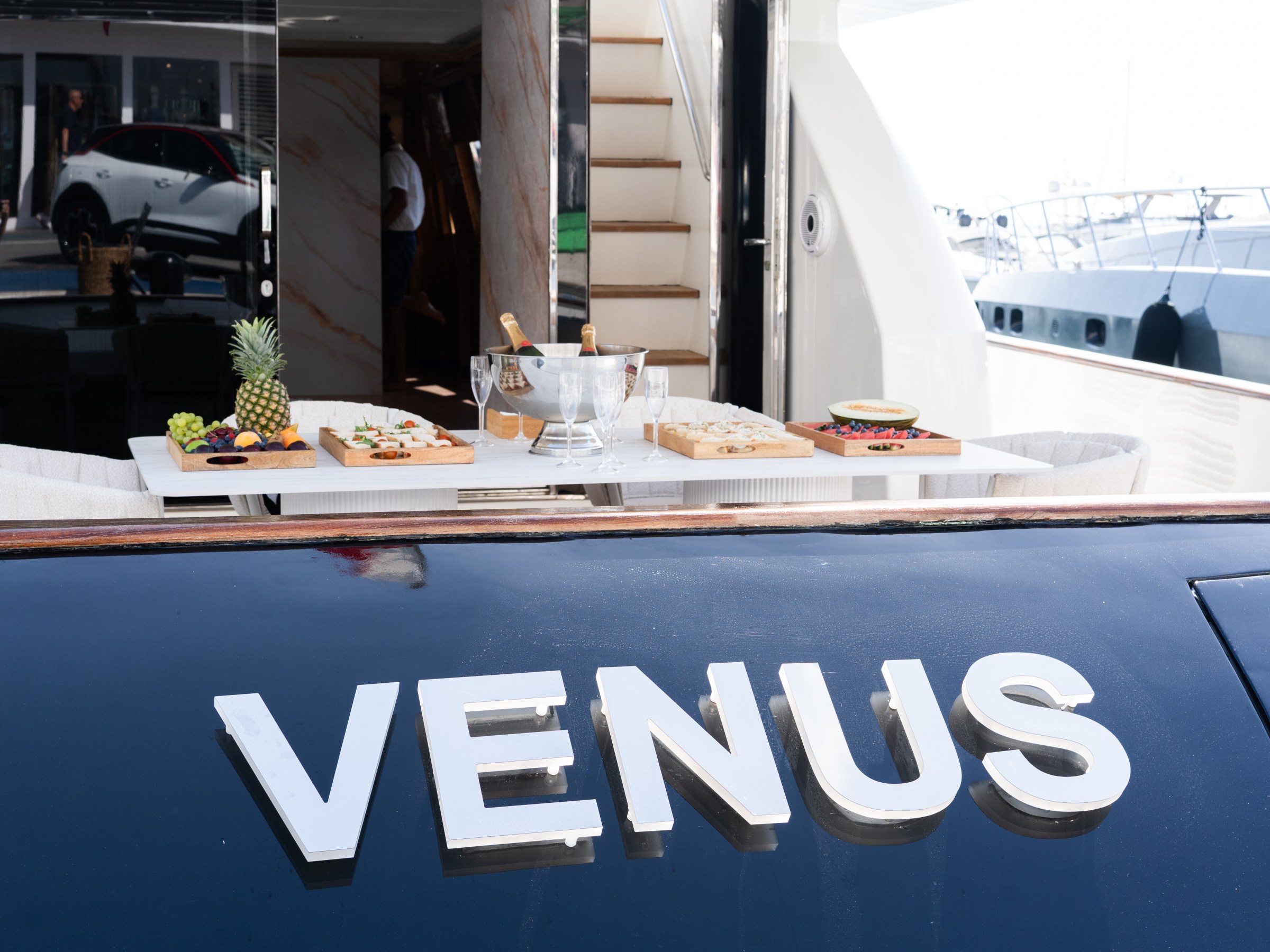 Back of a yacht named 'VENUS' with a table set for dining, featuring fruits and champagne.