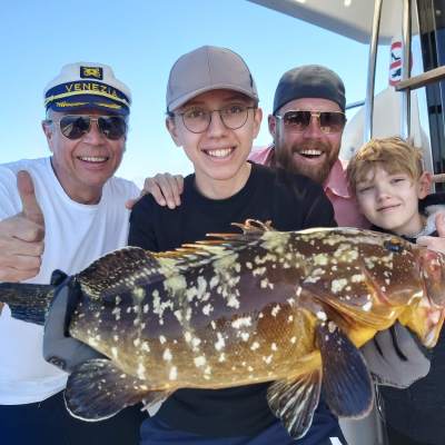 Four people smiling on a boat, holding a large fish and giving thumbs up.