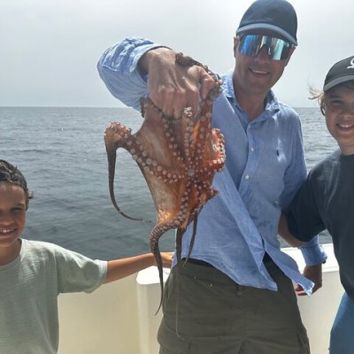 Three people on a boat, one holding a caught octopus, with ocean in the background.