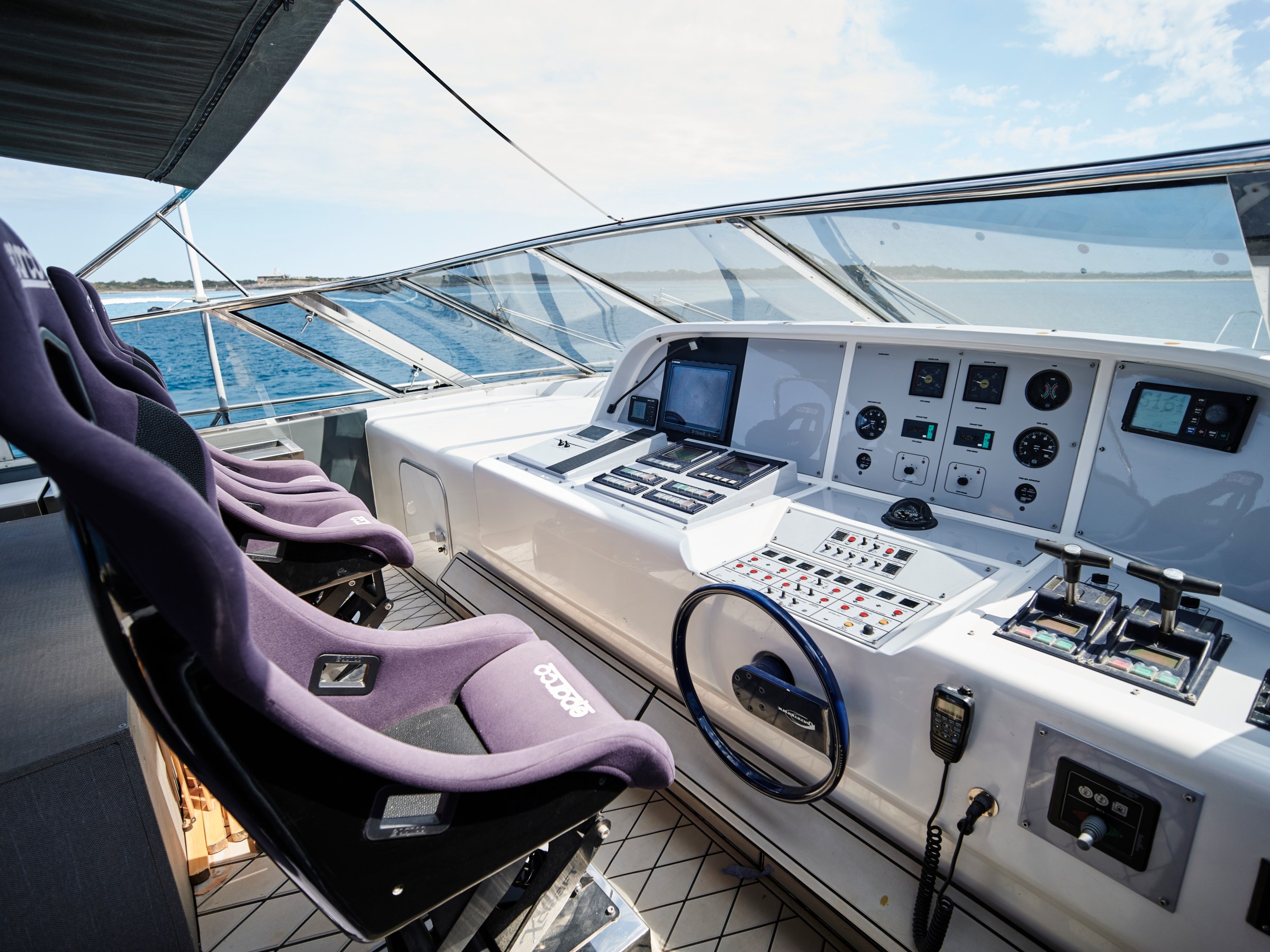 Boat cockpit with control panels, steering wheel, and four purple seats, facing the sea.
