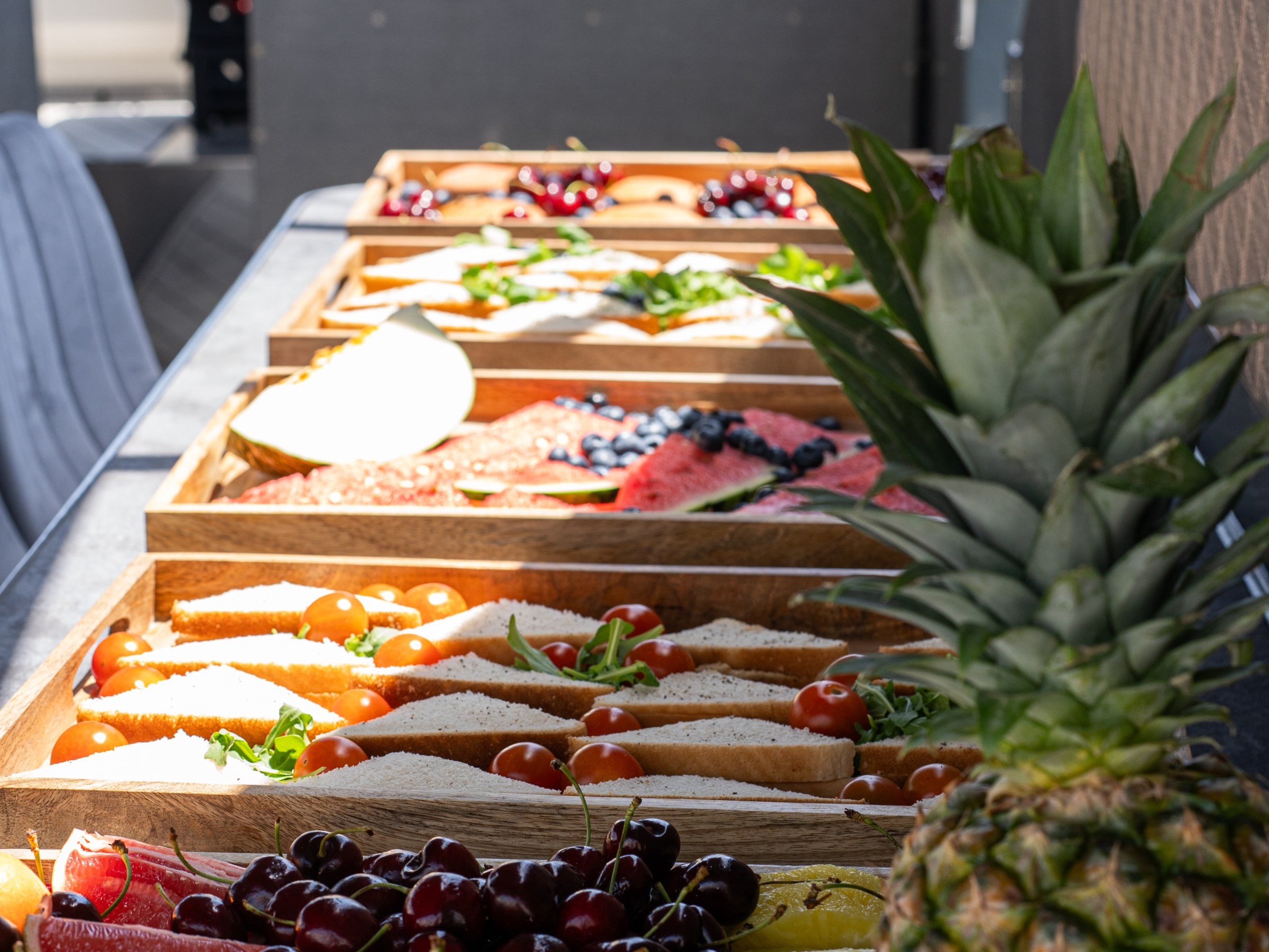 Tray of assorted fruits and sandwiches on a boat's table, including pineapple, cherries, and melons.