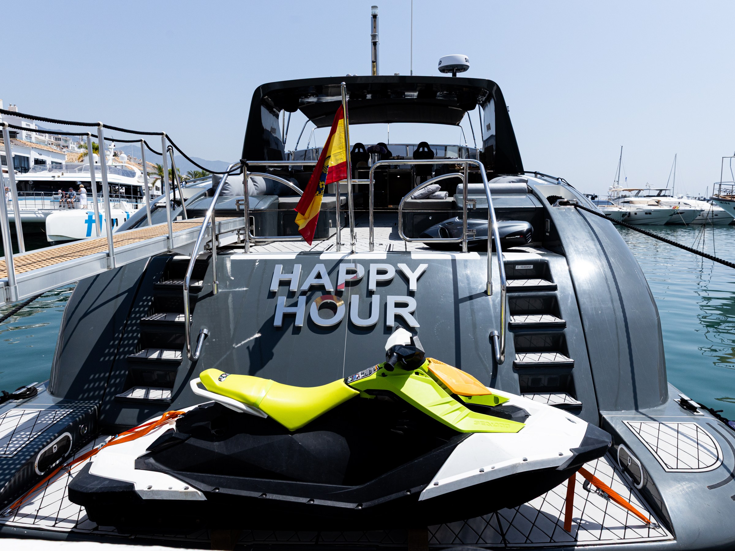 Rear view of a yacht named 'Happy Hour' with a Spanish flag and a jet ski on the deck.