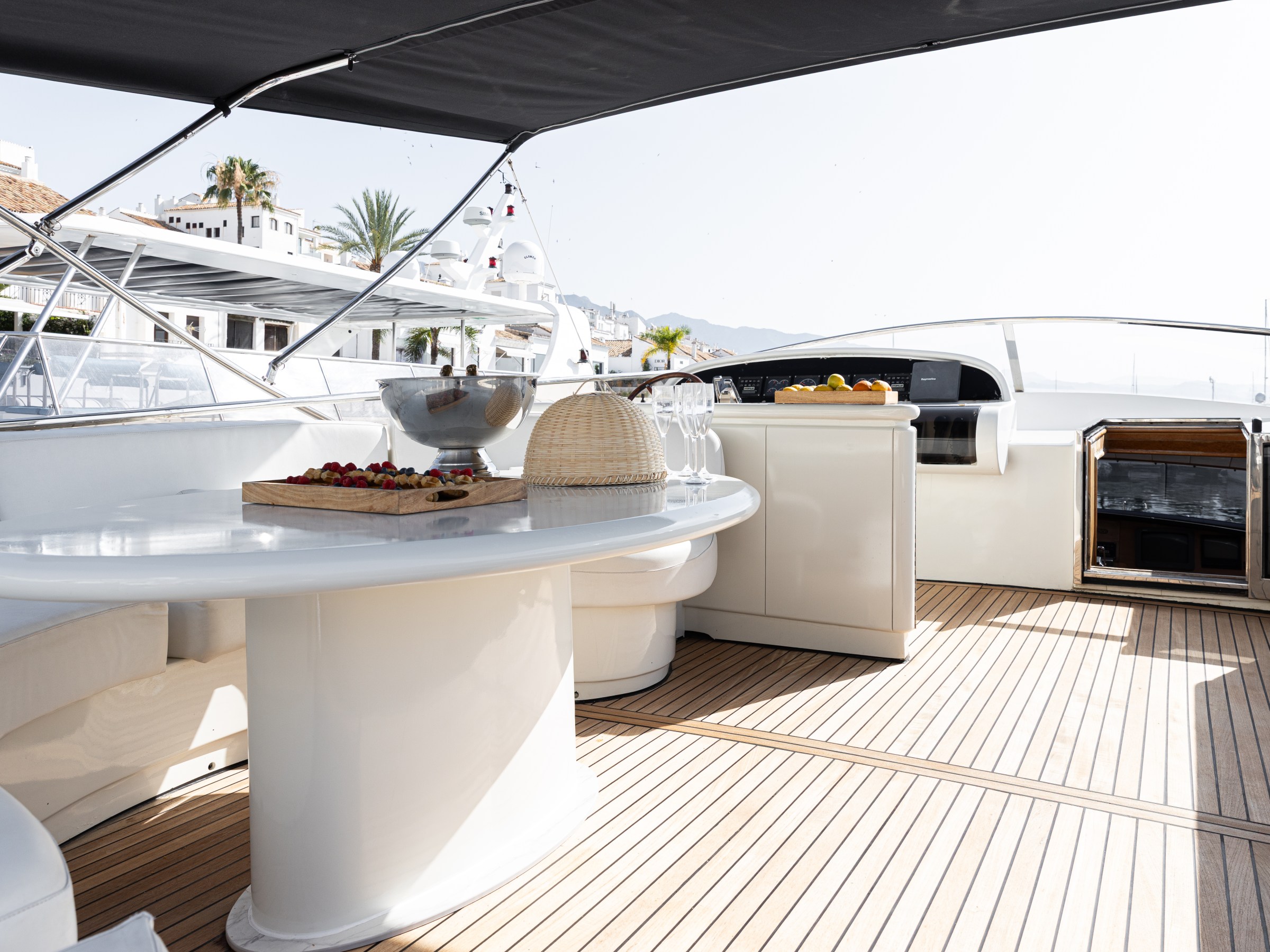 Luxury yacht deck with table, chairs, fruit, ice bucket, covered by canopy.