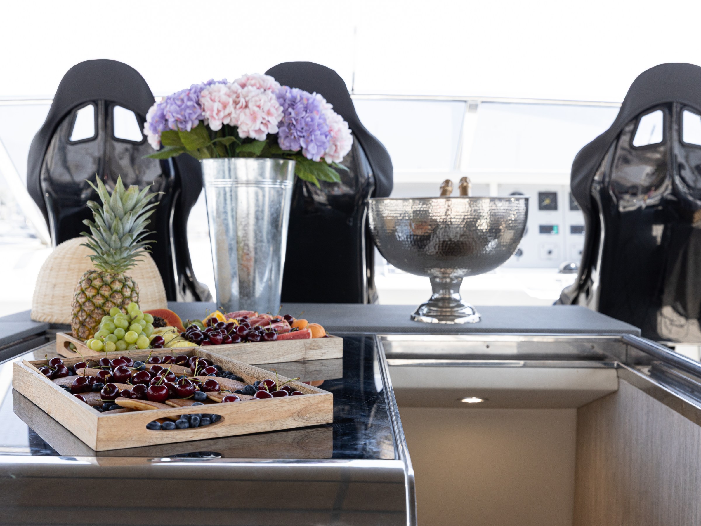Fruit trays, a pineapple, flowers, and an ice bucket on a yacht's deck with black seats.