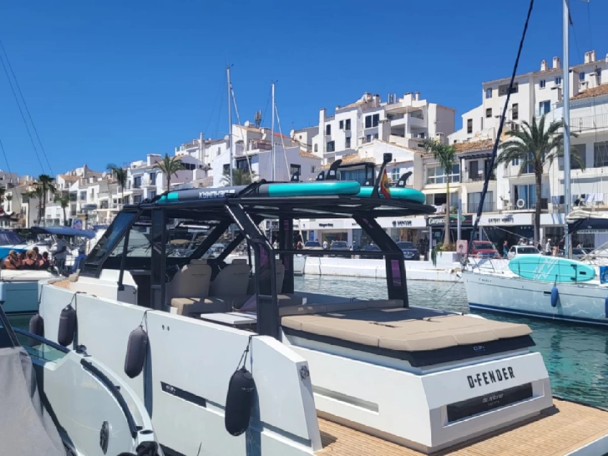 Boat docked in a marina with buildings and palm trees in the background on a sunny day.