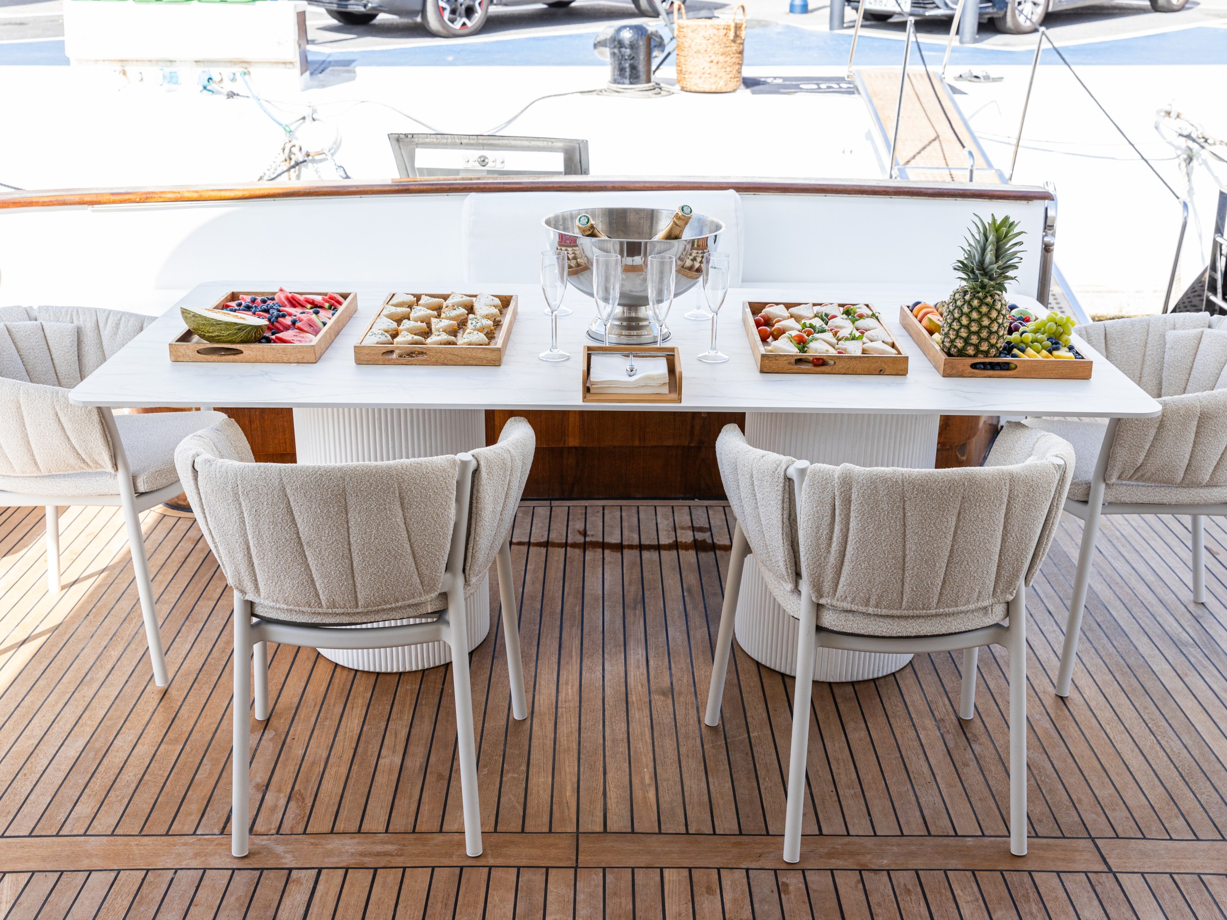 Boat deck table set with trays of fruit, snacks, and a champagne bucket, surrounded by plush chairs.