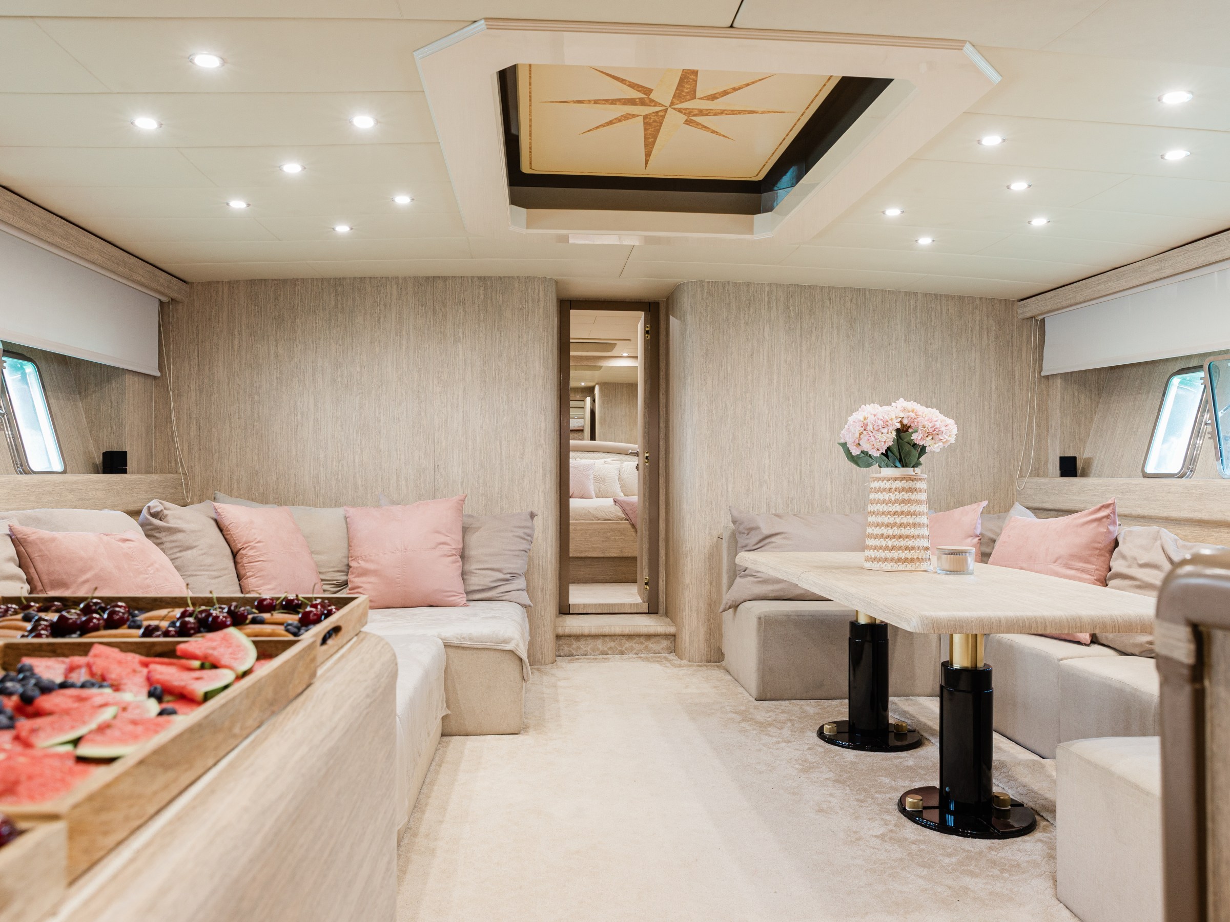 Luxurious yacht interior with beige seating, pink cushions, a dining table, and fruit platter.