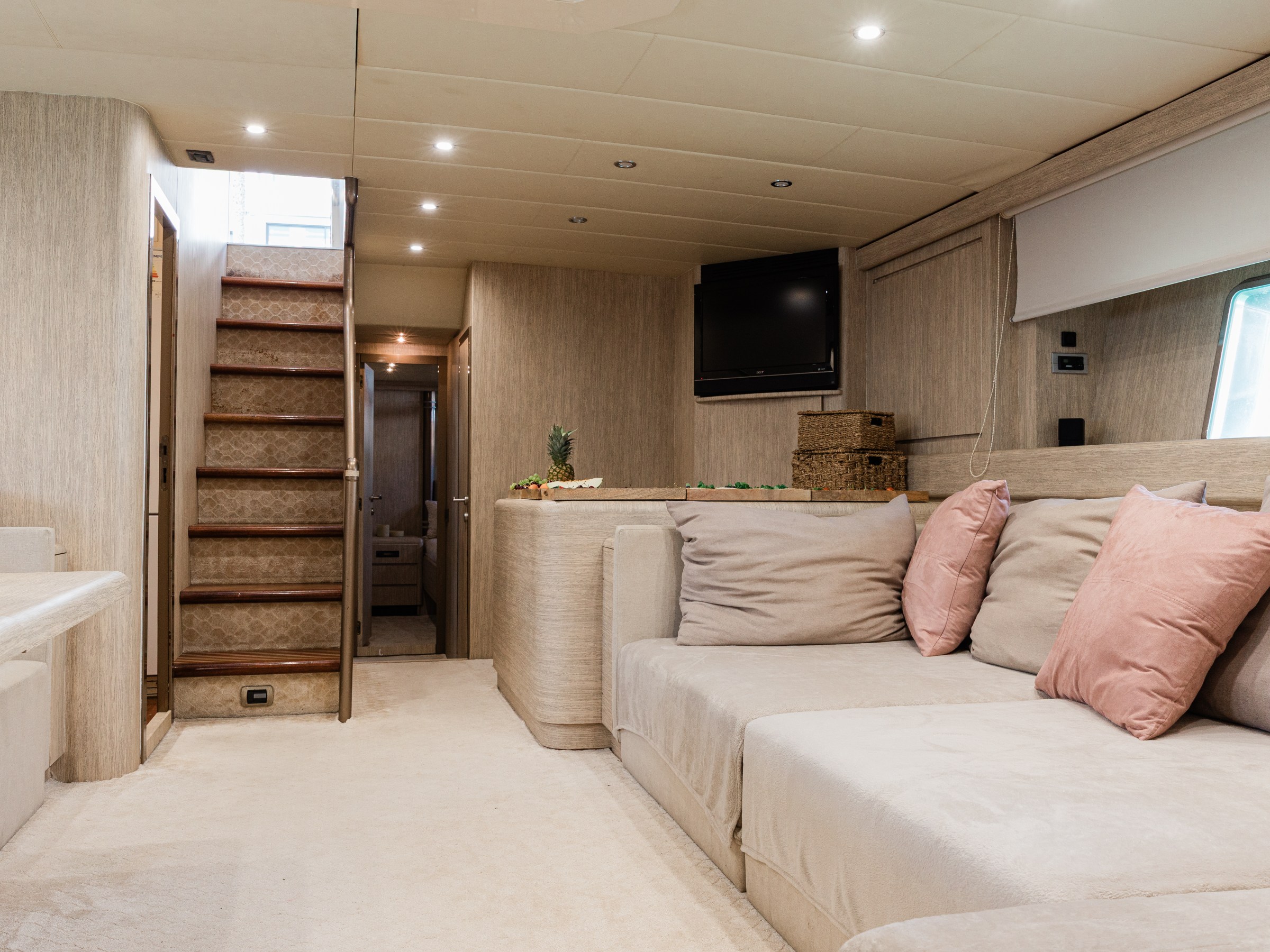Luxurious yacht interior with beige sofas, pink pillows, stairs, TV, and large windows.