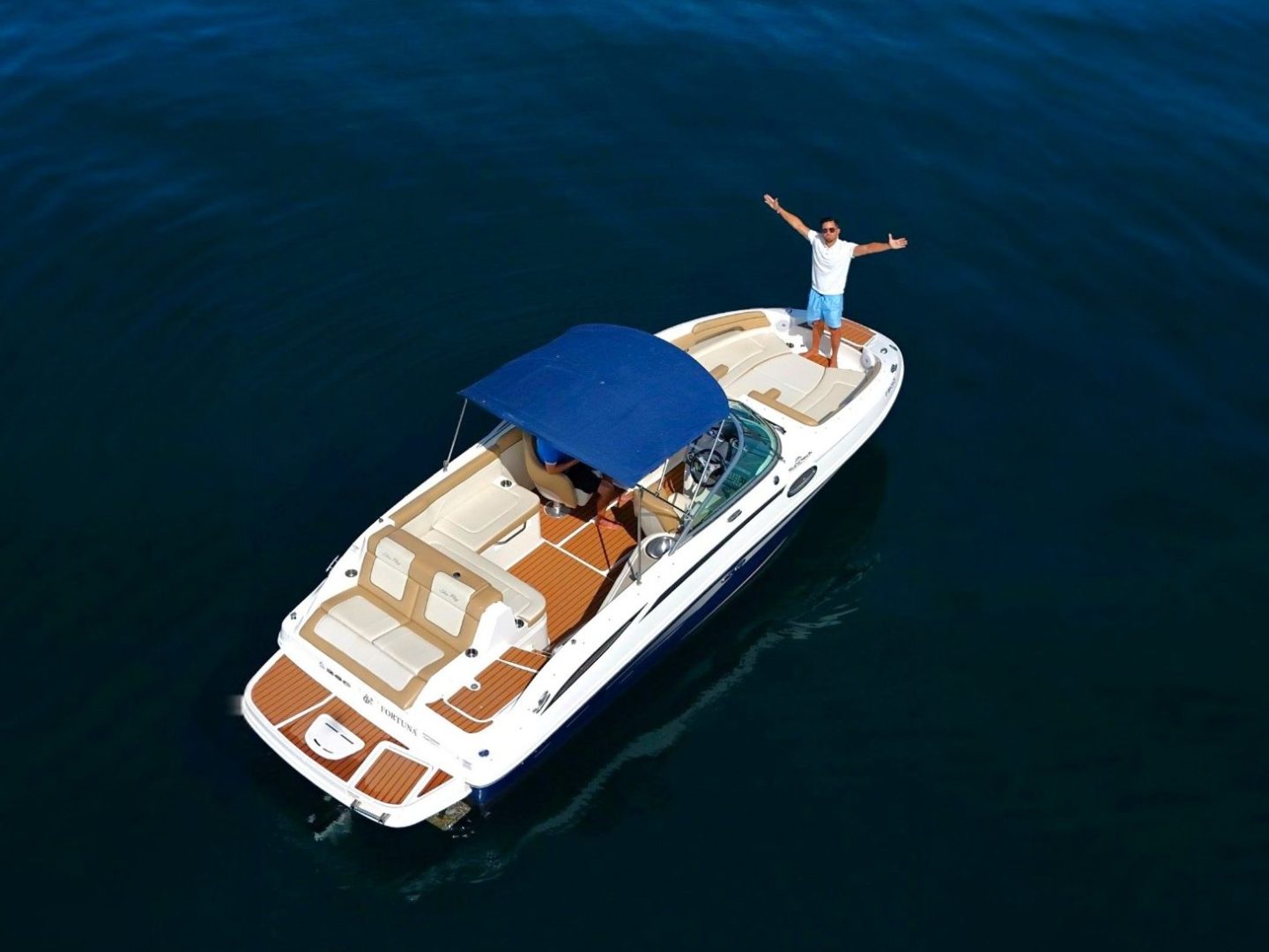 Aerial view of a person standing on a small motorboat in blue water, arms outstretched.