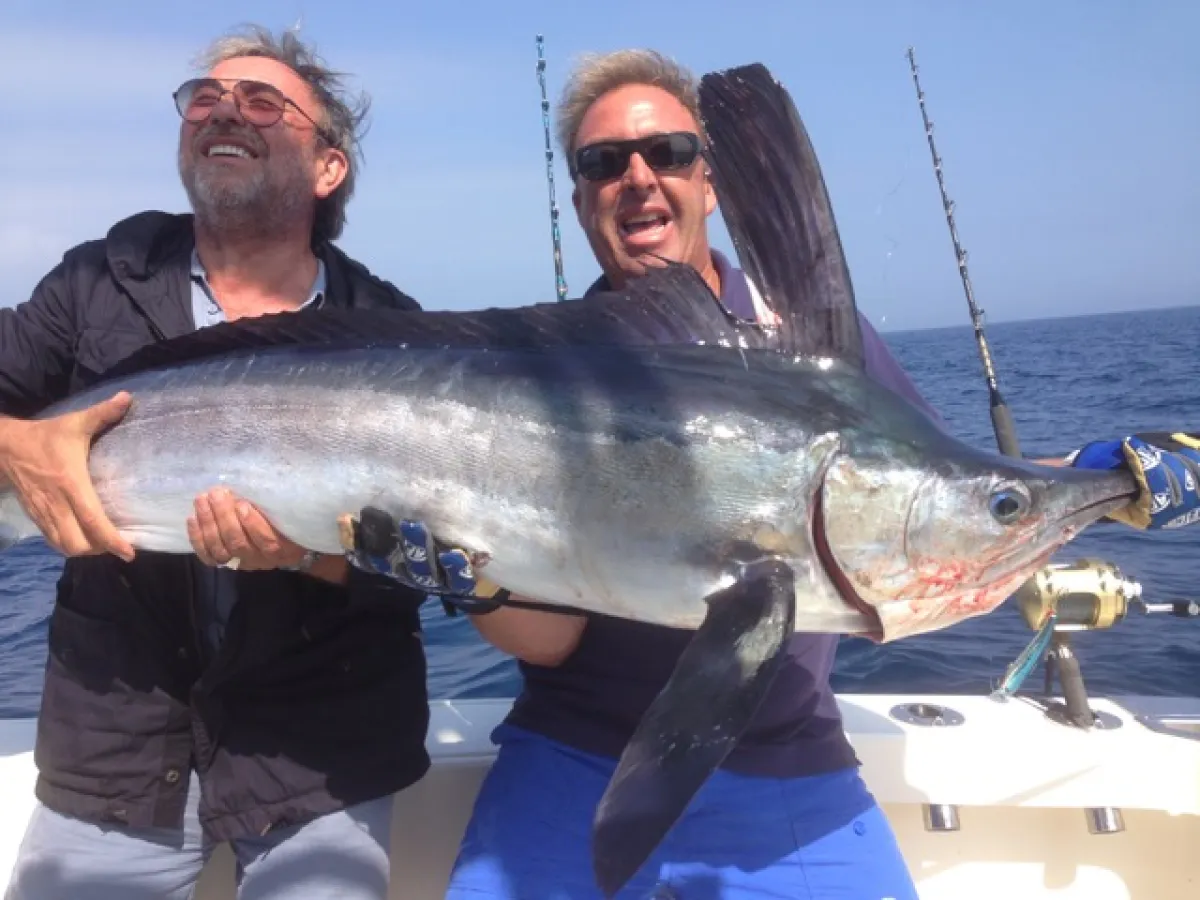 Two men on a boat holding a large fish with fishing rods in the background.