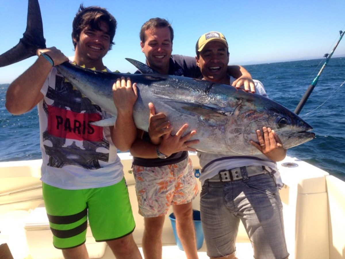 Three men on a boat holding a large fish against a backdrop of the ocean.