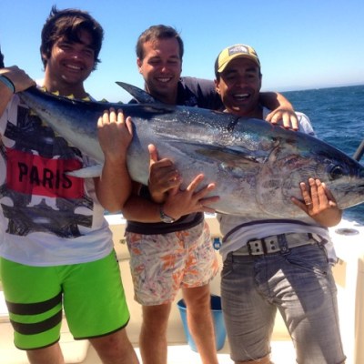 Three men on a boat holding a large fish against a backdrop of the ocean.