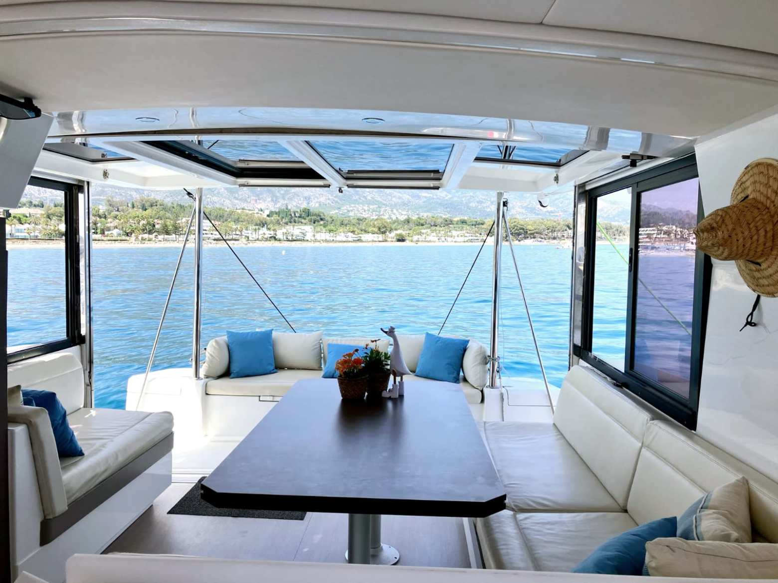 Inside view of a boat, showing seating area with cushions and table, overlooking calm water and distant shore.