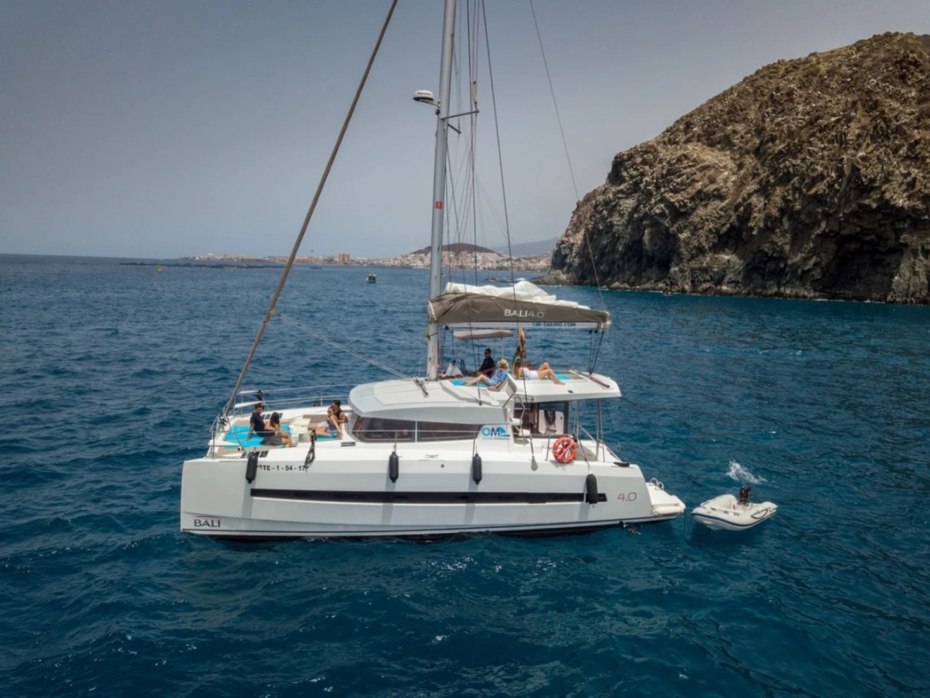 Catamaran with people on deck sails near rocky coastline in clear blue water.