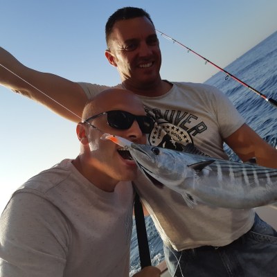 Two men on a boat, one is pretending to bite a caught fish, both smiling, with fishing rods visible in the background.