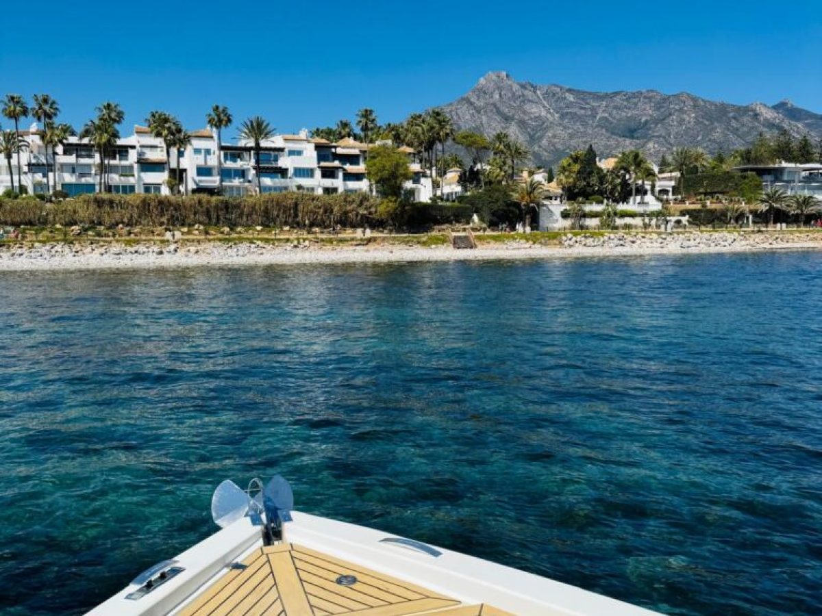 Boat bow facing coastline with palm trees, buildings, and mountains under a clear blue sky.