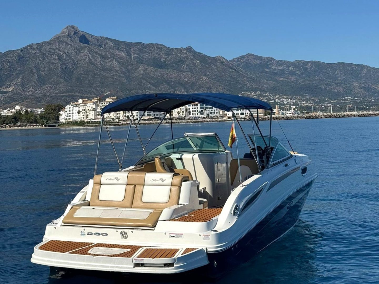 Boat on a calm sea with mountains and buildings in the background under a clear blue sky.