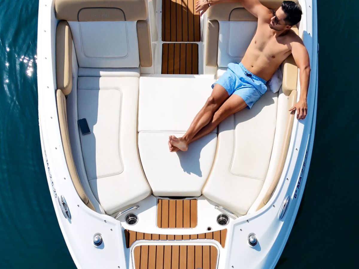 Man in shorts sunbathing on a yacht with white seating against a wooden deck on calm water.