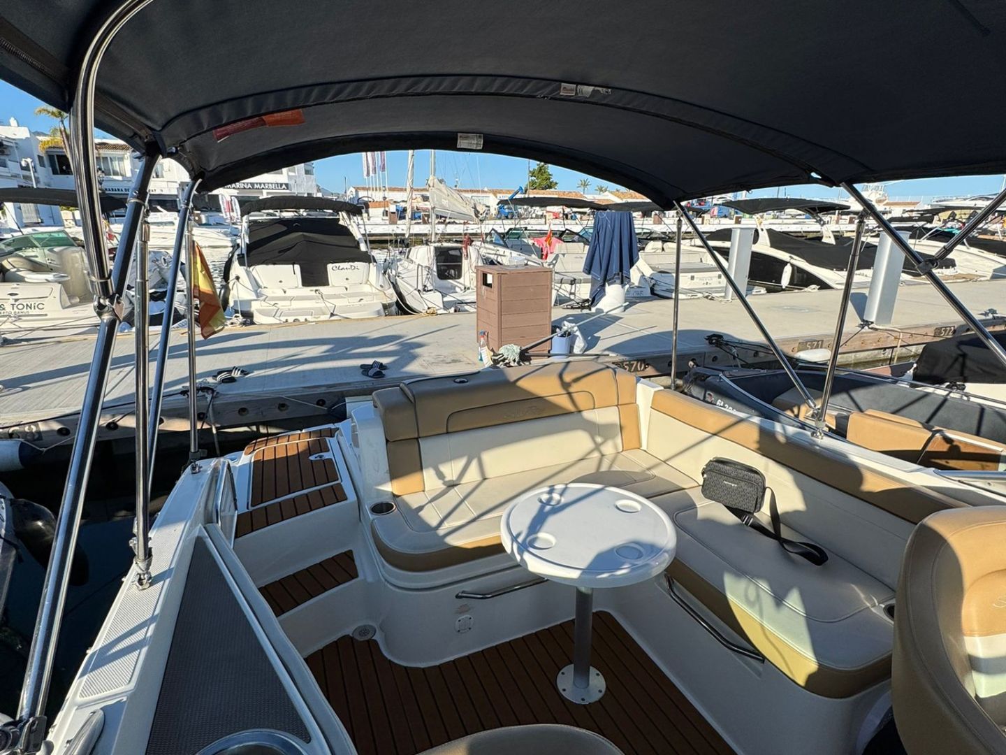Interior of a boat with tan seats, a round table, and a canopy overhead, docked at a marina.