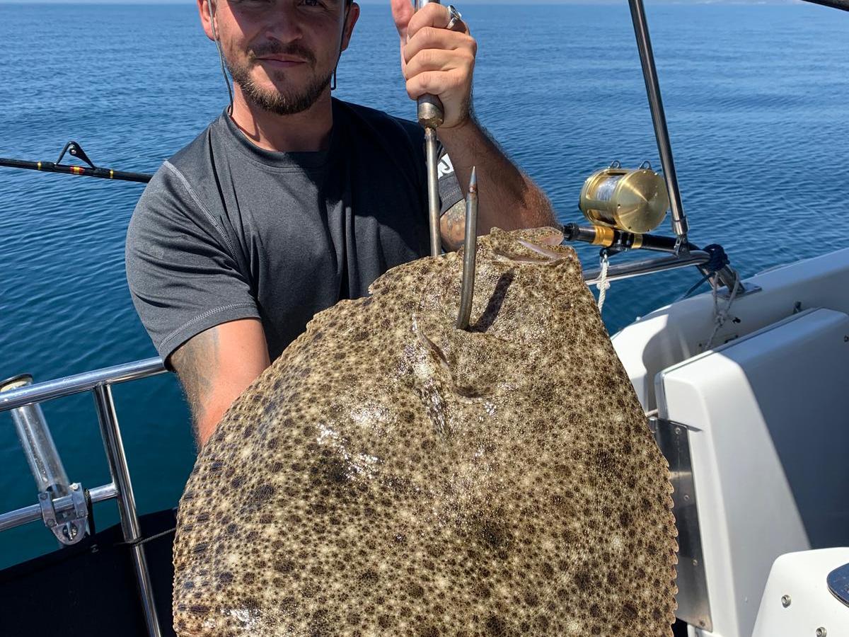 Person on a boat holding a large speckled flatfish with a blue ocean in the background.