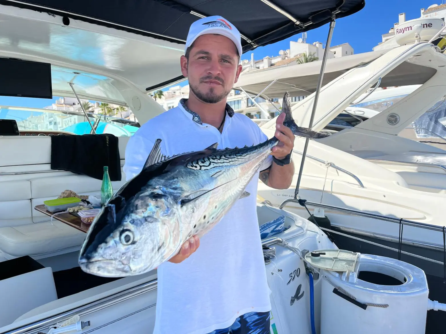 Person on a boat holding a large fish, with yacht interior and clear sky in the background.