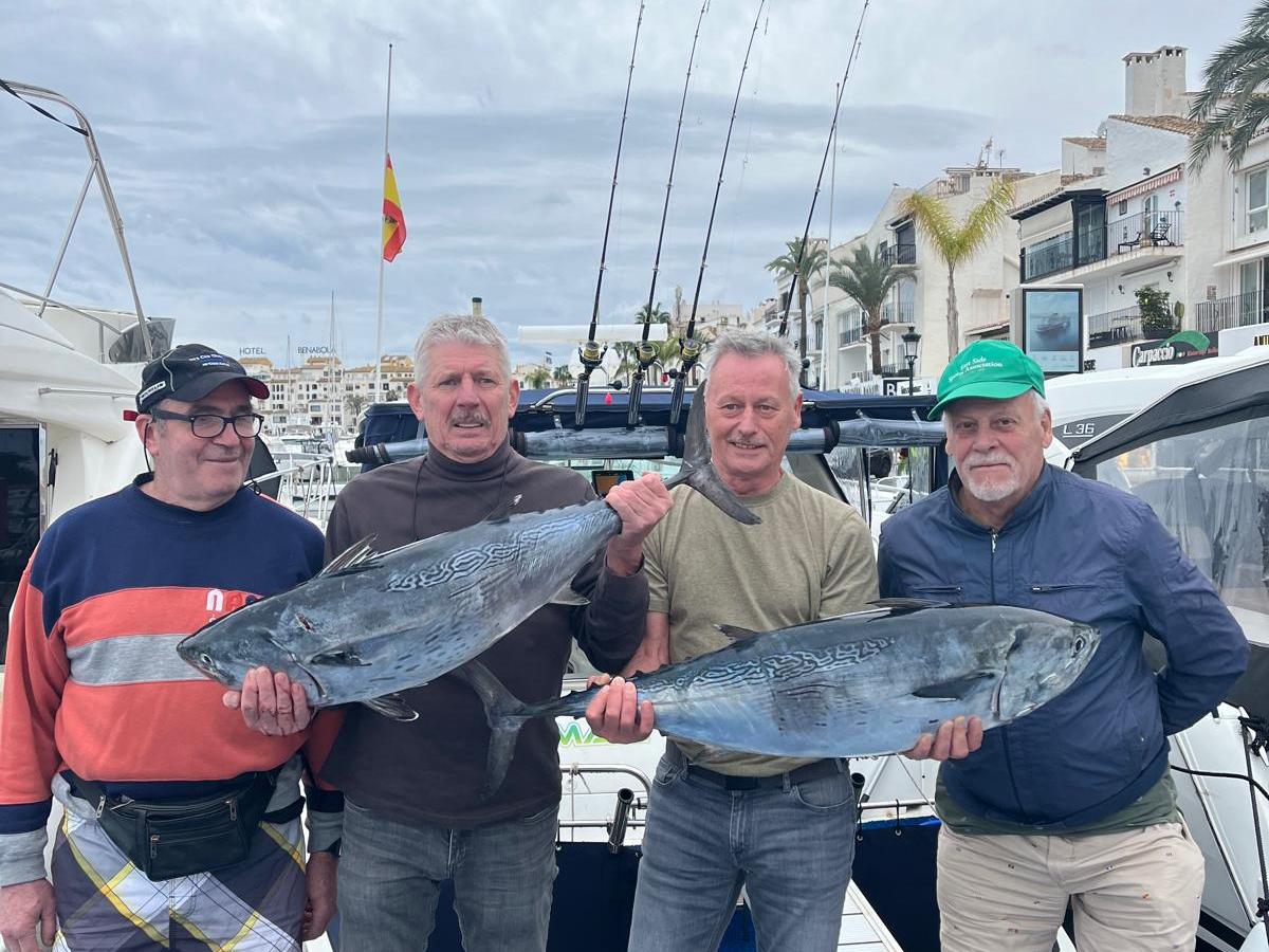 Four men on a dock holding large fish, with boats and buildings in the background.