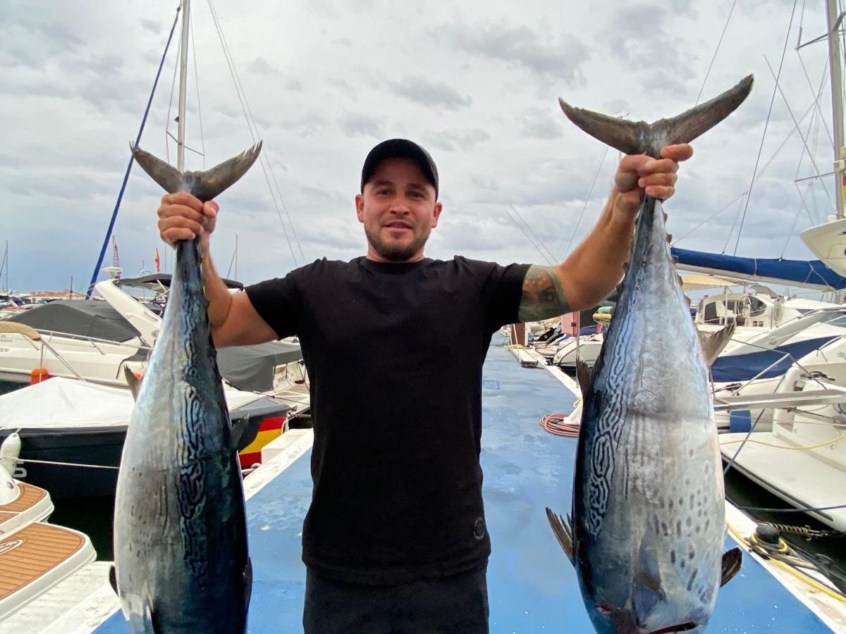 Man in black shirt holds two large fish at a marina, boats visible in the background.