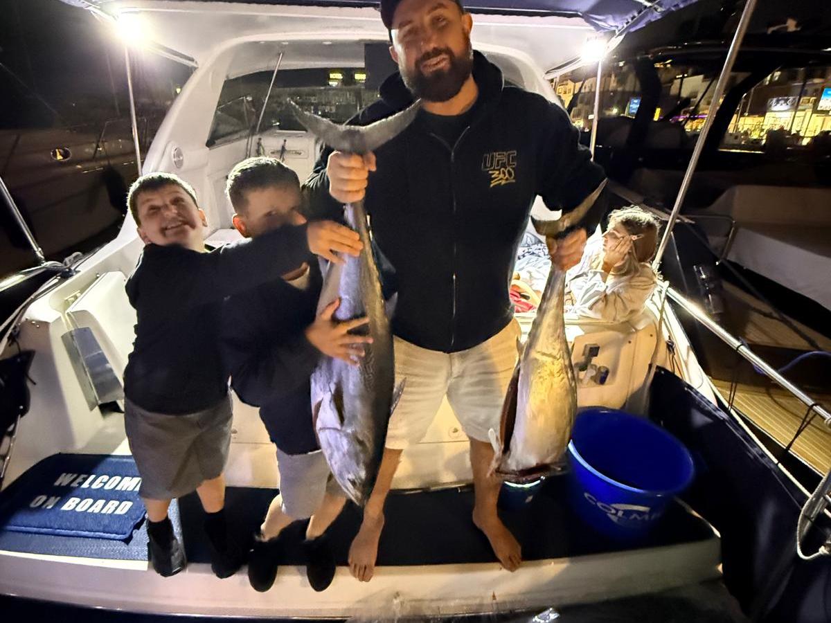 Man and two children smiling on a boat, holding large fish at night.
