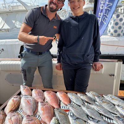 Two people smiling behind a table filled with a variety of fish near a marina.