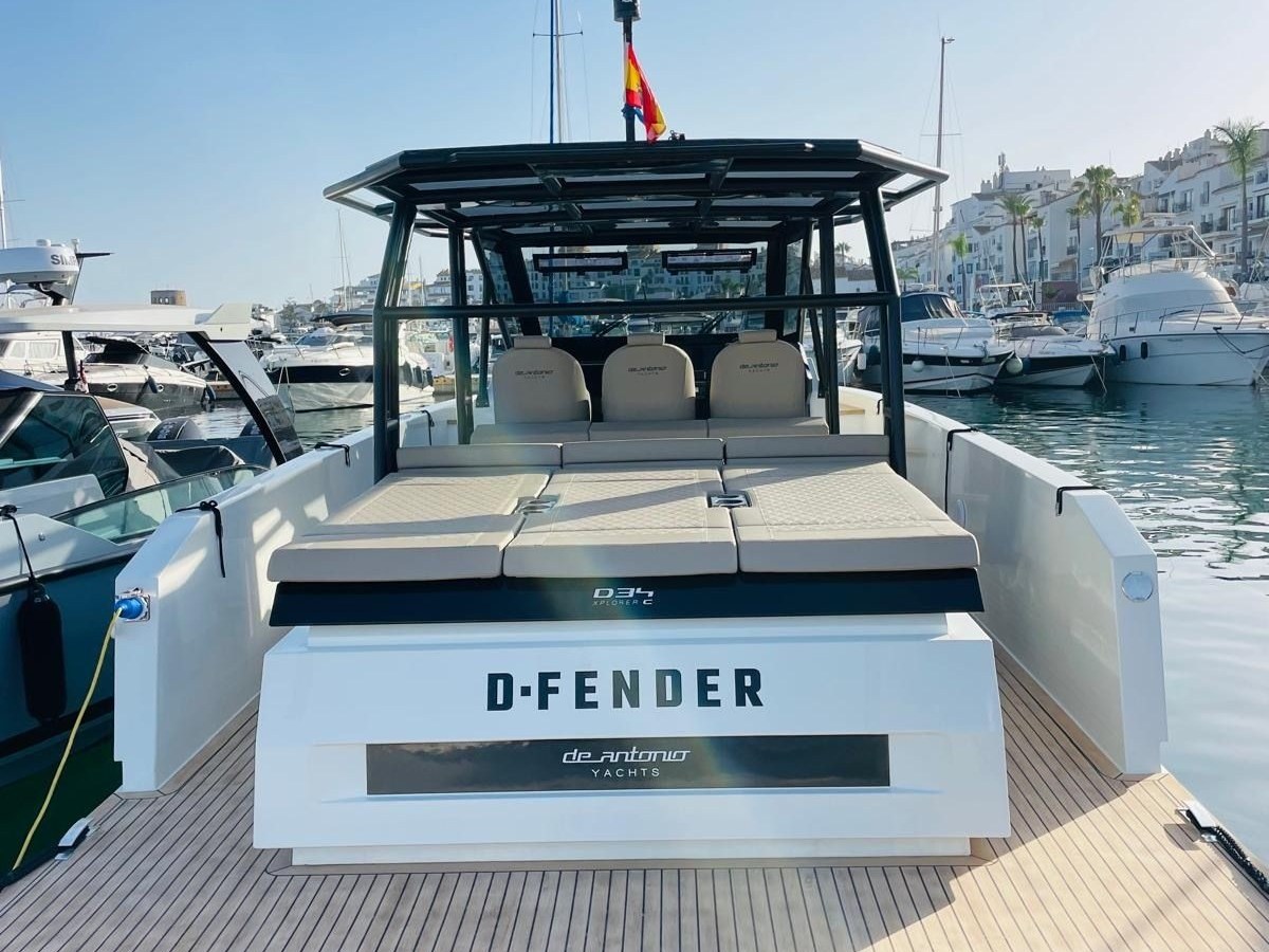 View of a modern yacht named D-Fender docked at a marina under a clear blue sky.