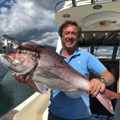 Man in blue shirt holding a large fish on a boat with cloudy sky and buildings in background.