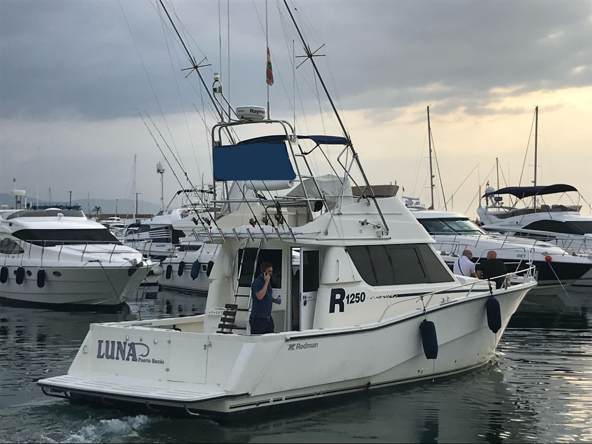 White motorboat with antennas in a marina, labeled 'Luna' with people onboard, under cloudy sky.
