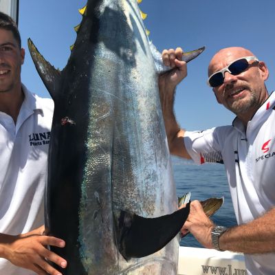 Two men on a boat holding a large fish with shiny scales against a clear blue sky.