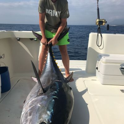 Person in green shorts and sunglasses holds large tuna fish on a boat.