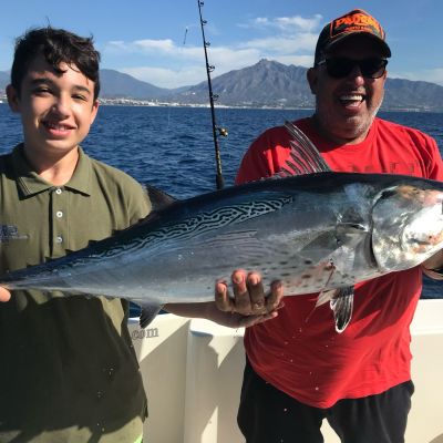 Two people on a boat holding a large fish with the sea and mountains in the background.