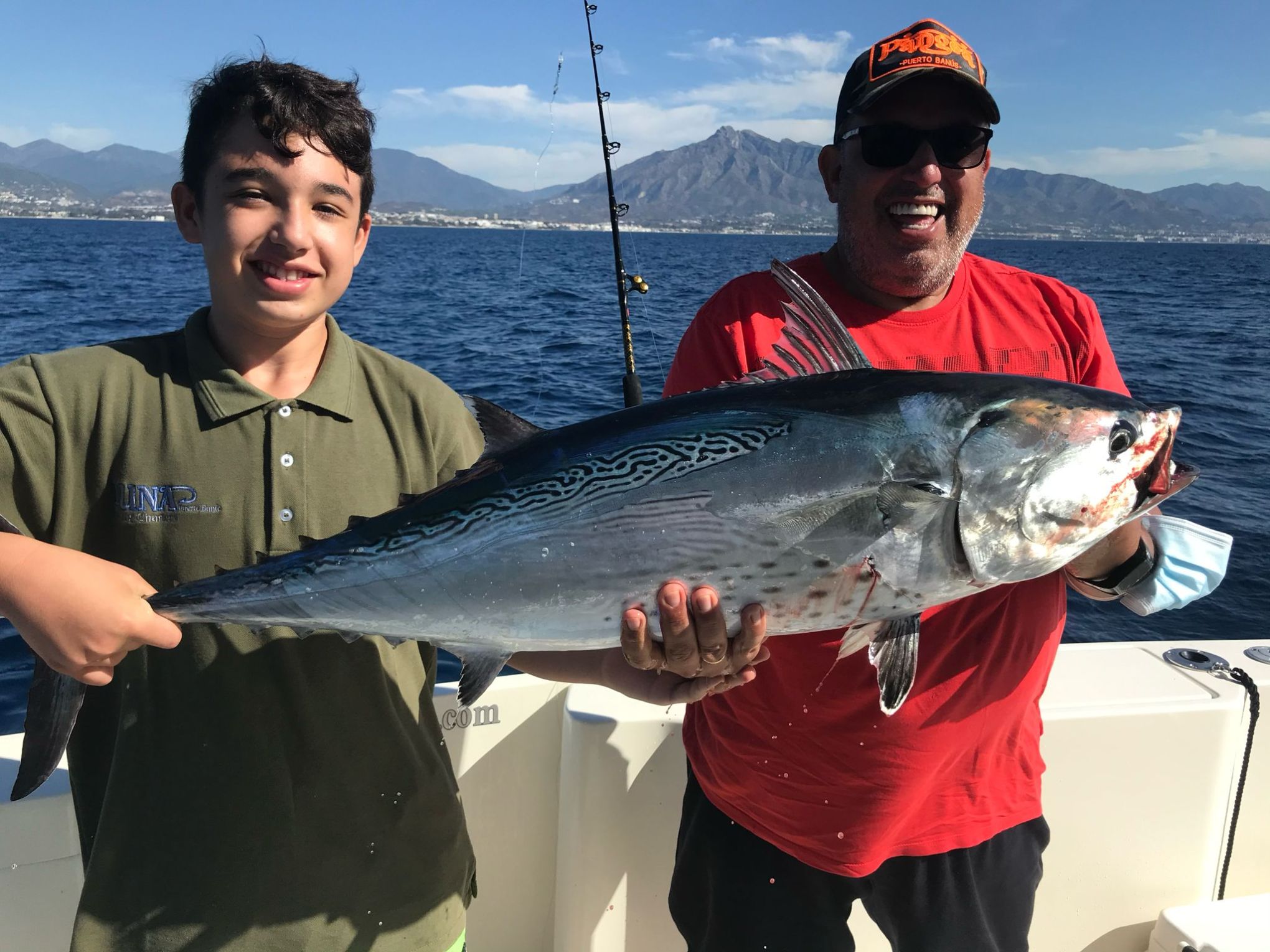 Two fishermen smiling on a boat holding a large fish with mountains and ocean in the background.