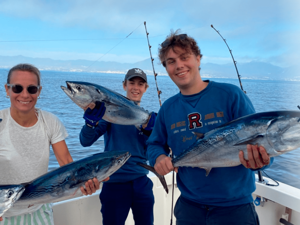 Three people on a boat holding freshly caught fish, with ocean and mountains in the background.