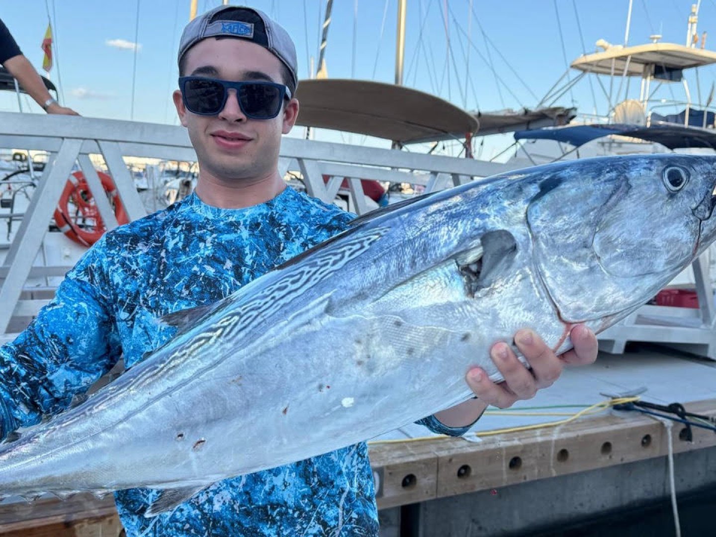 Person in blue shirt holding a large fish on a dock with boats in the background.