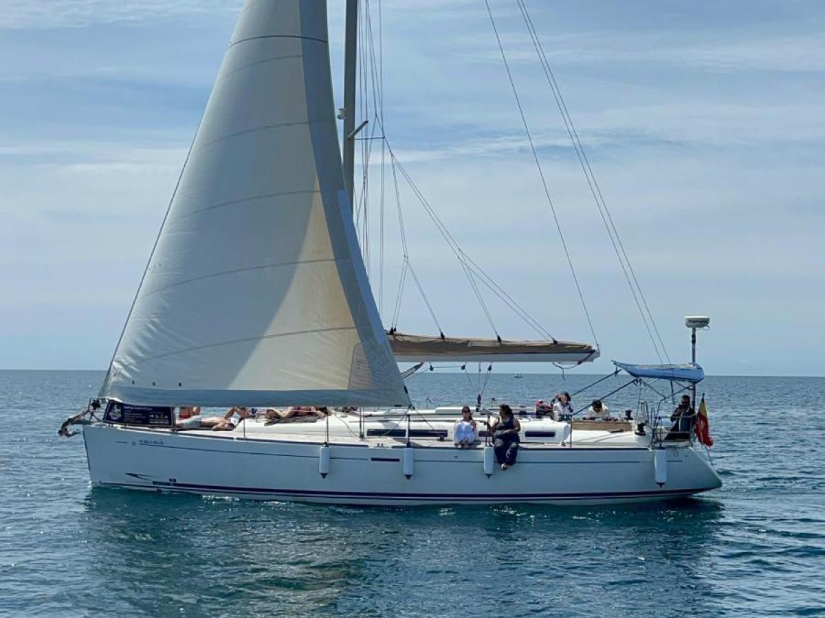 A sailboat with white sails gliding on clear blue water under a partly cloudy sky.