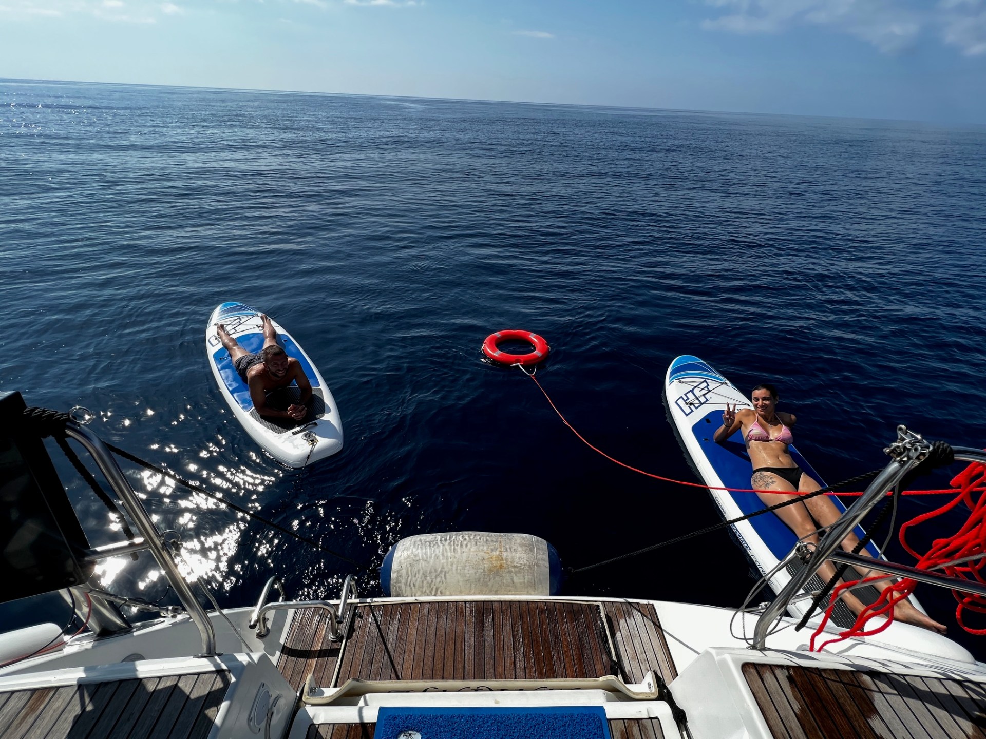 Two people on paddleboards beside a boat on calm water with a red lifebuoy between them.