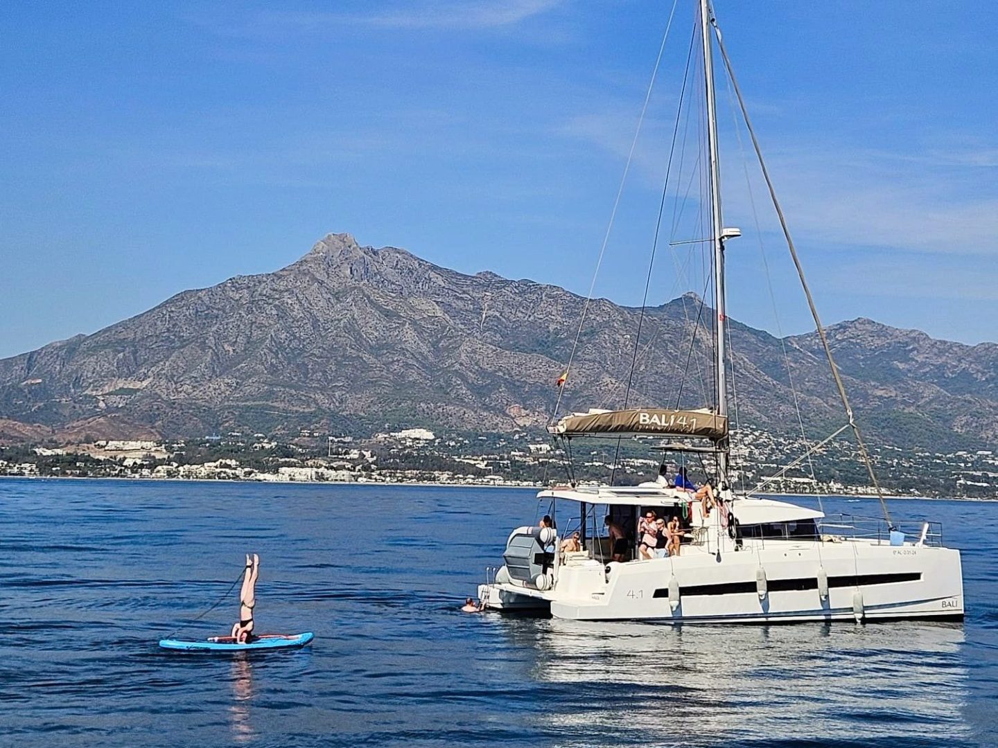 Sailboat on calm sea with mountains in background, person doing headstand on paddleboard.