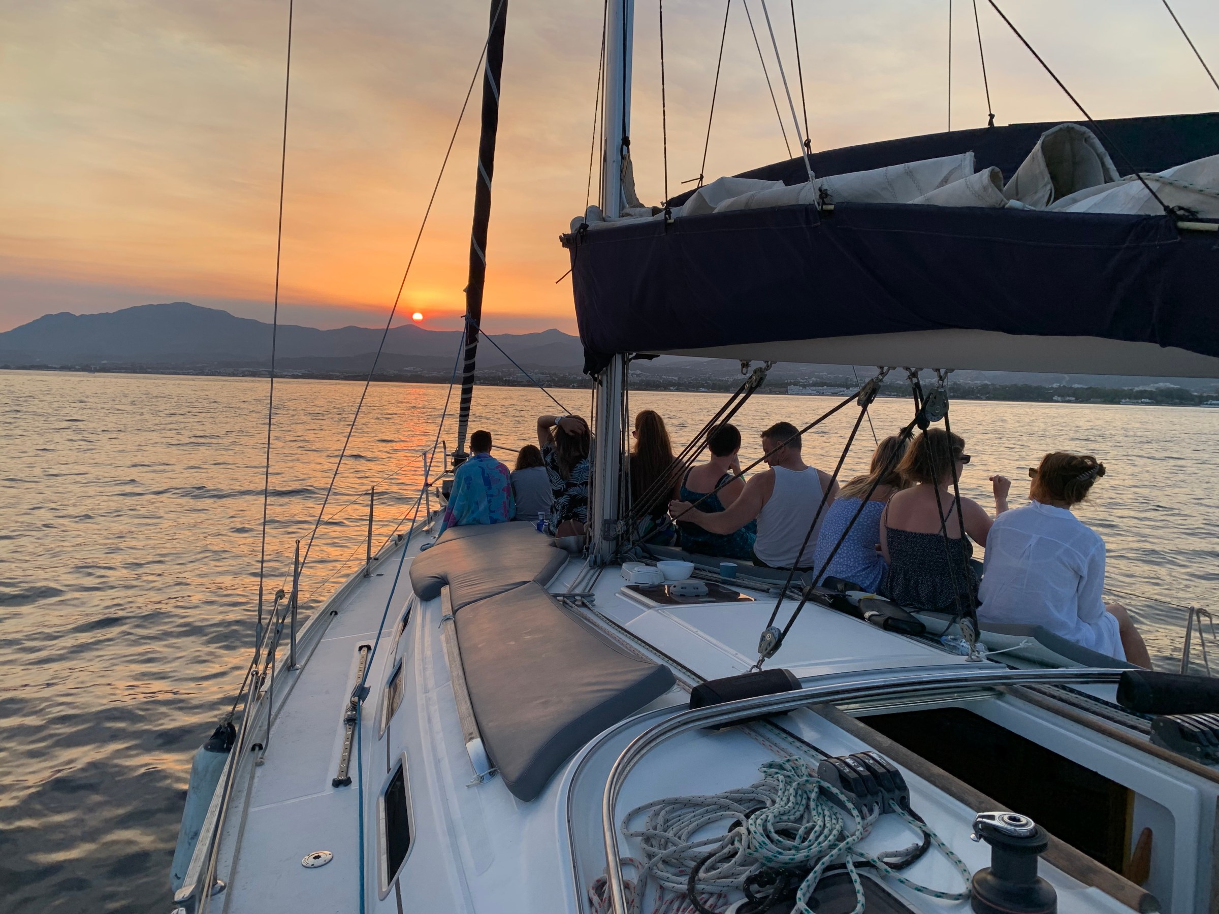 People on a sailboat watching the sunset over the ocean with mountains in the background.