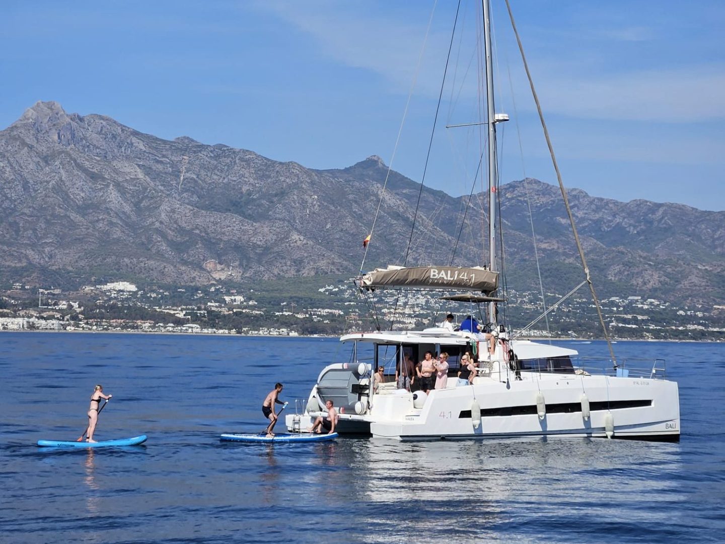 Catamaran on calm sea with people paddleboarding nearby, mountains and clear sky in background.