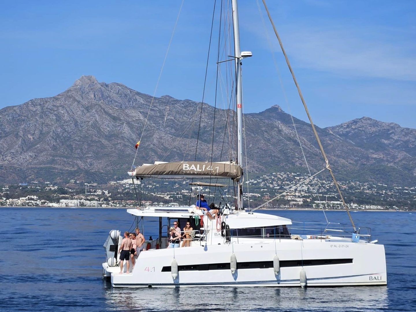 Catamaran with people on board sailing near a mountainous coastline under a clear blue sky.