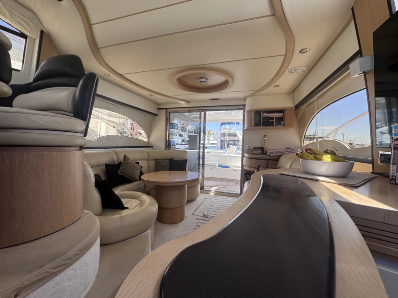 Luxurious yacht interior with sofas, table, and kitchen area, facing marina view.
