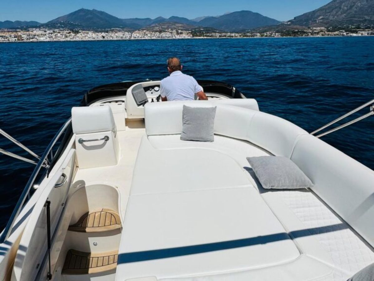 Person steering a boat with cushioned deck, blue sea and distant mountains under clear sky.