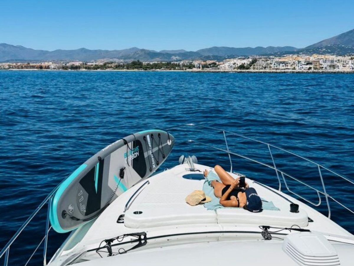 Person sunbathing on a boat with paddleboards, overlooking a coastal town and mountains.